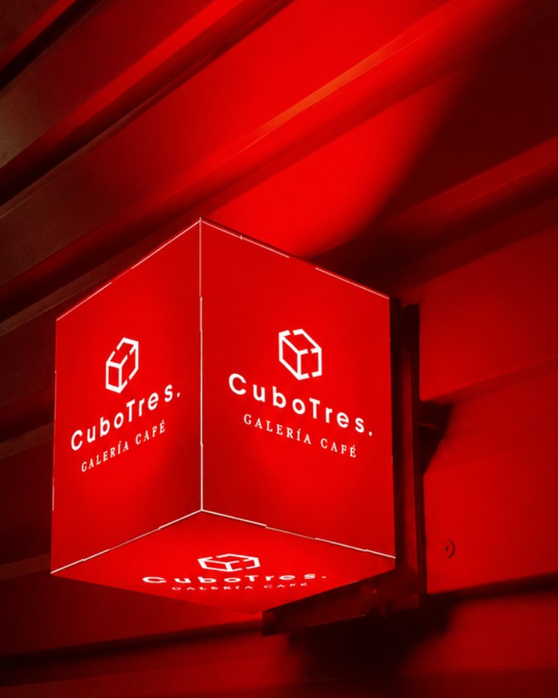 Cubo luminoso de tres caras con el logotipo de CuboTres Galería Café, encendido en rojo sobre la fachada del local.