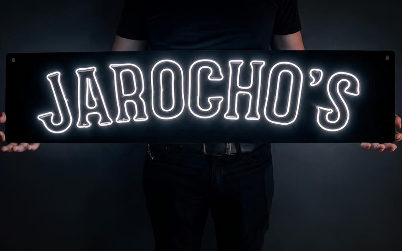Letrero de neón blanco con la palabra Jarocho's montado sobre una placa negra, sostenido con las dos manos.