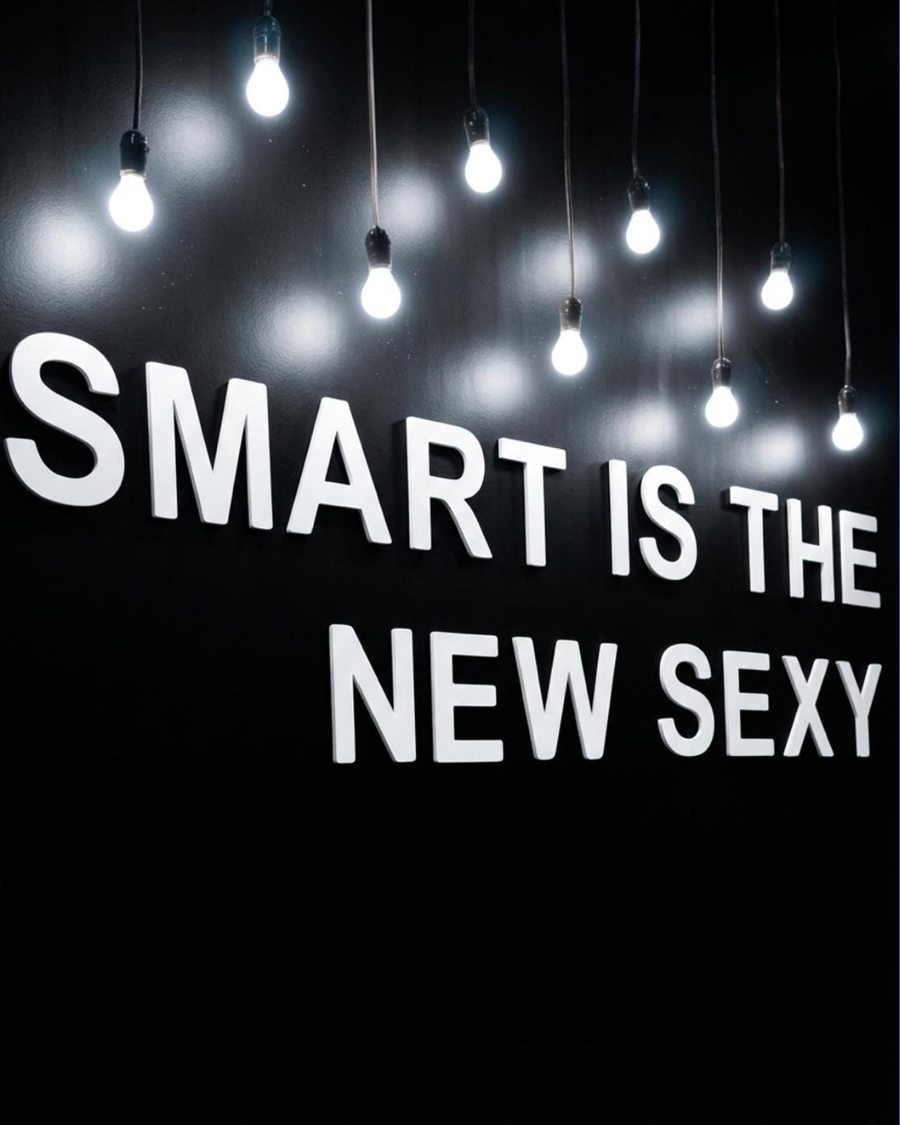Letras corpóreas blancas con la frase Smart is the new sexy sobre un muro negro, iluminadas por focos colgantes.