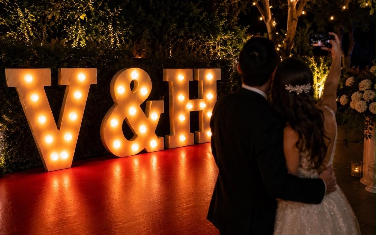 Letras gigantes V y H con foco tipo marquesina encendidas en el jardín de una boda, con los novios de espaldas frente a ellas.